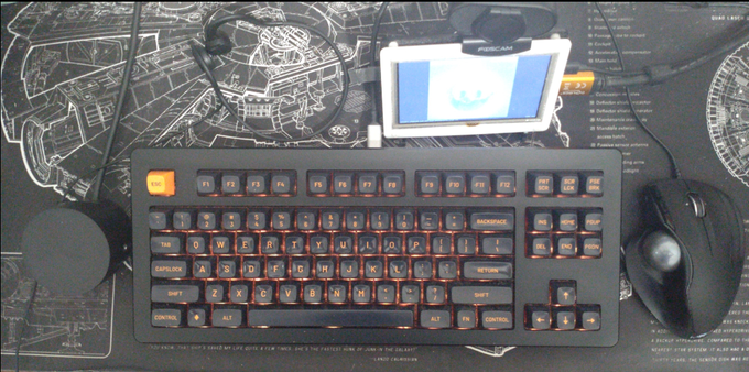 A black Rama U80-A Seq. 2 ten-key-less (TKL) mechanical keyboard with orange-on-black Skiidata keycaps in tall MT3 profile and black braided USB-C cable with matte silver plug sitting on a desk mat with a white-on-black deskmat depicting a StarWars Millenium Falcon blueprint. The spacebar has noticeable shine from usage with the right thumb.

Other visible devices:

Right a dark grey, ergonomic, thumb-operated, wired Mod-IT trackball with glittery medium grey ball.

Left the large, black volume knob from a (not visible) Logitech Z533 speaker set.

Behind the keyboard right of the keyboard's USB cable there is a little 7" screen with a white and black acrylic case + stand and a webcam sitting on top of it. The screen shows the cover of Jean-Michel Jarre's album Oxygene and has an orange-metallic HDMI plug coming from the right side.

Left of the cable there is a black AfterShockz wired bone conduction headset laying on the deskmat.