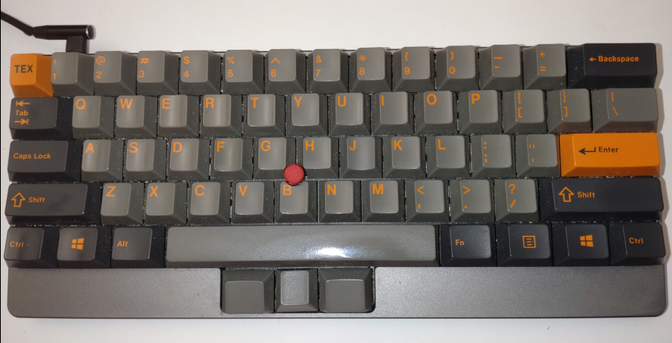 A Tex Yoda II 60% keyboard with a red thumbstick (well-known from ThinkPads) and an orange-on-medium-and-dark-grey Cherry profile keycap set sitting on a white desk. It additionally has three medium grey blank keys in front of the spacebar which function as mouse button. The outer two mouse keys are inclined towards the user. The spacebar has clearly visible shine from usage with the right thumb.