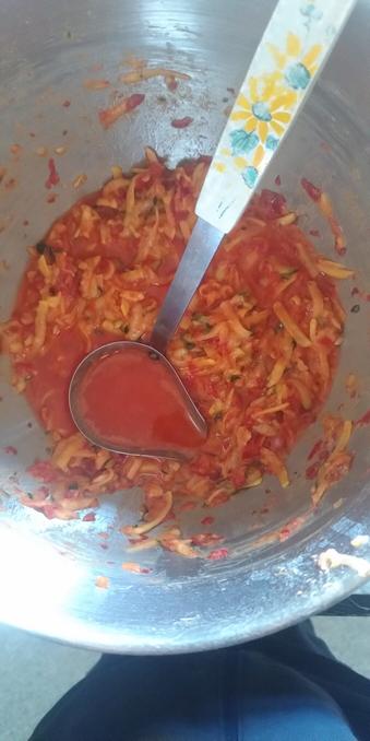 Eine kleine Schöpfkelle über einer Masse aus geraspelter Paprika, Zucchini, Zwiebel, Knoblauch, Kartoffel in einer Edelstahlschüssel.