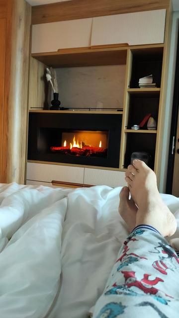 An artificial log fire burning & spluttering away on embedded in an open cupboard structure with the bed end & the poster's foot in the foreground