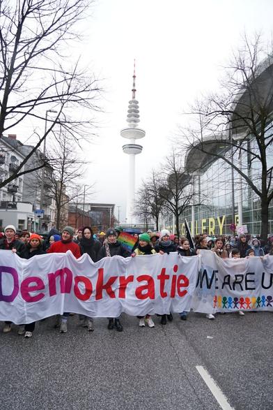 Menschen mit Mützen und Handschuhen halten das Frontbanner mit der Aufschrift "Demokratie WE ARE UNSTOPPABLE". Zentral im Hintergrund ist der Fernsehturm, rechts die Messe.