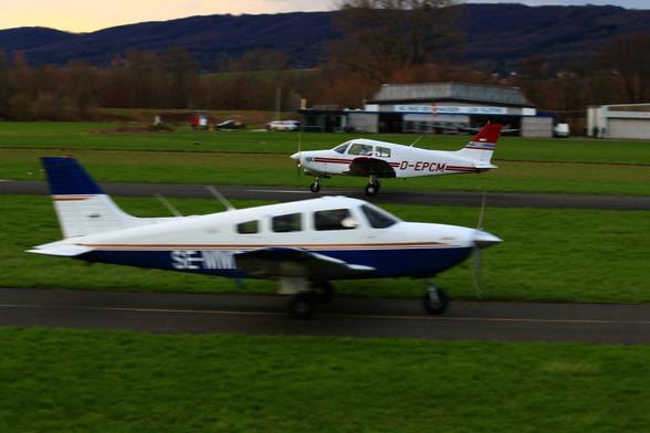 Two low-wing light aircraft, one on the runway, white with with red stripes, taking off, one blue/white on the taxiway in the foreground with some motion blur.