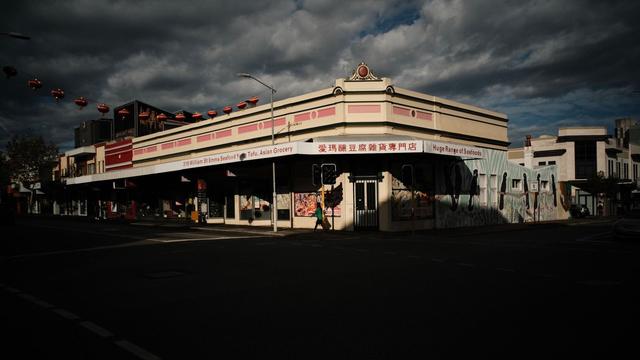 Sunlight brightens a corner Asian food store in an urban area. An individual walks around the corner in front of the store. The foreground is in a shadow. The sky is partly cloudy.