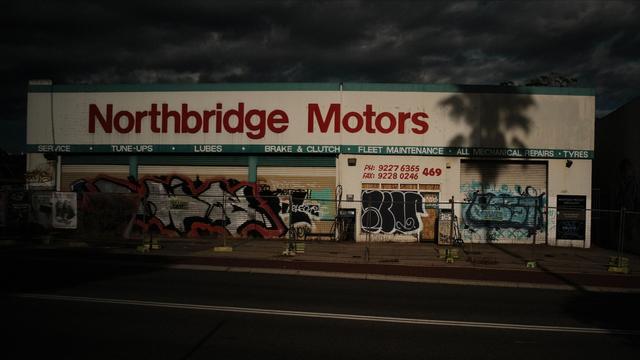 Sunlight brightens a closed automotive shop in an urban area. A palm tree shadow falls on the right side of the shop. A chain link fence runs the length of the area in front.