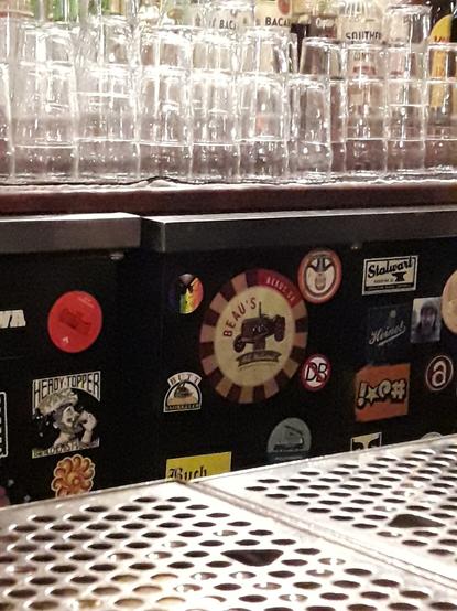 A round sticker with a rainbow flag and Lainey's Luck logo - a hand drawn cat - is seen on a wall of stickers in a pub