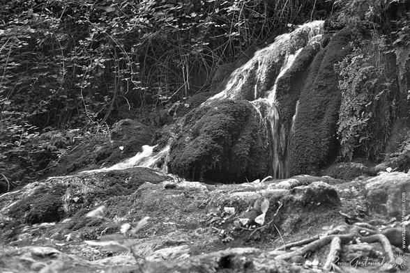 A black-and-white photo of a small stream cascading down large mossy boulders.