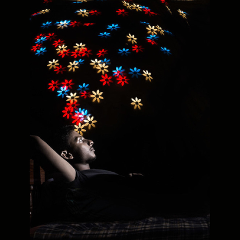Daydream by Maximilian Imran Faleel on Flickr: photograph of a young black man in a black t-shirt against a black background, leaning back and gazing up, with colorful illustrated daisy-like flowers in red, yellow, and blue floating up from his head