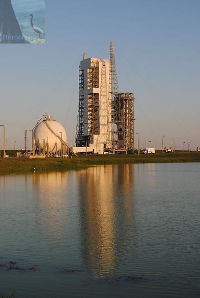 A vertically-oriented photograph taken outdoors and in the evening, of a white tower on the other side of a pond. On the left side of the white tower and a gray, truss tower is on the right.
