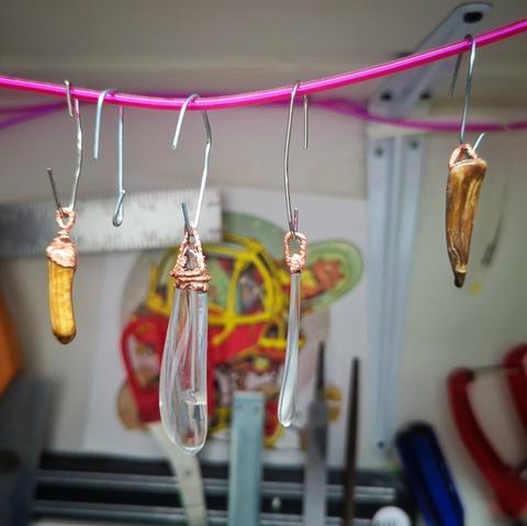 A pink cord has 5 hooks hanging on it. The first hook on the left holds a copper covered brown stone (petrified wood) about 1.5 inches long. The next hook is empty. A 2 inch piece of glass with copper wound around the top. A very thin piece of glass with only a slight bit of copper at the top. Final hook contains a canine tooth shaped piece of petrified wood  with a copper top.