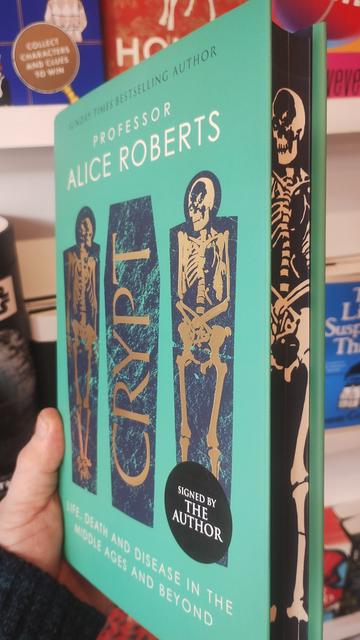 Angled side on view of Crypt by Alice Roberts, to show the cover, with golden foiled skeletons in their tombs, and the side of the book with "spredges" (sprayed edges), done in black with a white skeleton image