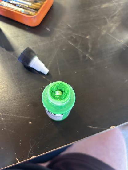 An open green paint with its cap off on a black surface, and part of an orange container in the background.
