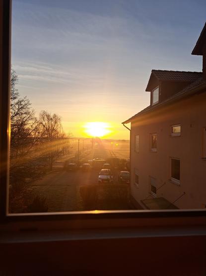 Ansicht aufgehenden Sonne. Gelber Horizont. Links Bäume,  Mitte Parkplatz mit Utos und dahinter Feld, rechts Hausfassade.