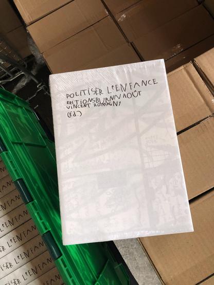 Photo d'un exemplaire de "Politiser l'enfance" sous blister, vu en plongée, sur une pile de cartons. On voit dans l'angle la gauche un bac norme Europe vert contenant d'autres exemplaires sous blister.