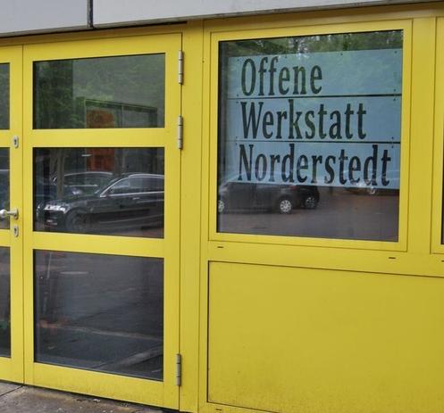 Eingang zur Offenen Werkstatt Norderstedt. Glastür mit gelben Rahmen. Schild mit Schriftzug Offene Werkstatt Norderstedt hängt daneben im Fenster. Die Wand und der Fensterrahmen sind ebenfalls gelb.