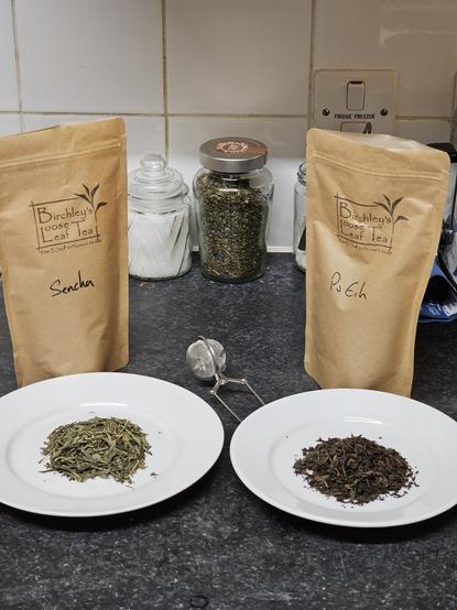 A photo of a basic tea set-up, including a peanut butter jar of peppermint, and two bags, one containing 普洱茶 Pu'er tea and the other containing Sencha. A couple of plates hold examples of the respective teas.