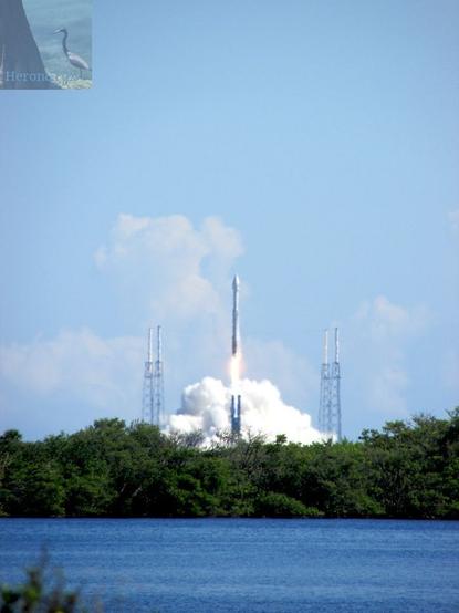 An outdoor, daylight photograph of a rocket launching. There is a body of water and strip of green brush in between the launch pad and the camera. The rocket is ascending on a column of orange flame. White smoke and dusk is being blown into a cloud behind the pad.