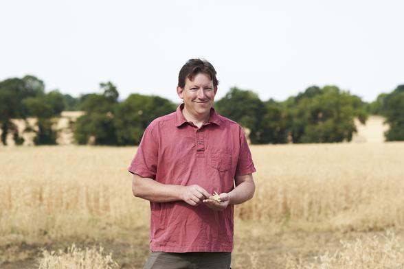 Josiah Meldrum, a man in a light red shirt, stands in a field of grains. Behind him are trees.