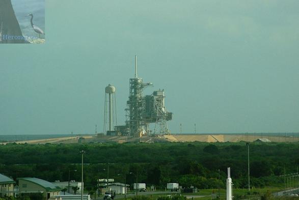 An outdoor, daylight photograph of a truss structure, including a tower near the center of the frame with a large, complex metal structure supported by the gray tower on the right. There is a water tower on the left.