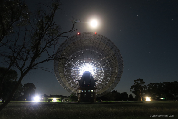 A large radio telescope, seen from behind and tilted towards the sky. Above it hangs the bright full Moon. There are several trees around as well.