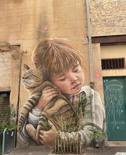 Streetartwall. The enchanting mural of a little girl with her cat was sprayed/painted on the outside wall of a single-storey old building in a narrow alley. In the style of a pastel drawing, you can see the brown-haired child with closed eyes, lovingly holding a brown and black striped cat in her arms and hugging it. A finely drawn picture with a lovely warm atmosphere. The photo shows plant stems fenced in by a low wooden fence in front of the mural and a green door with jellyfish graffiti next to it.
Info: The mural shows the homeowner's granddaughter with her cat Lulu. The mural is located in an alley of old buildings full of original graffiti and murals. The town of Séte is world-famous for its street art festival.