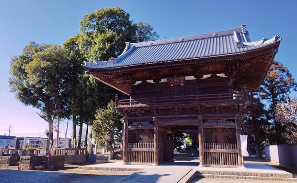 A large gate the size of a two-storey building made of wood that's withstood the test of time and weather thus far. It is framed by trees to either side and a cemetery on the left as well. The gate itself is placed more to the left as if it and the trees all formed a unit that had to be centered.