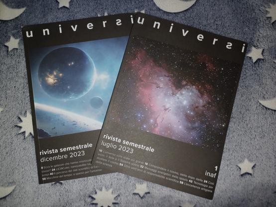 Due riviste appoggiate su una coperta grigia a fantasia di lune e stelle. Le riviste hanno una copertina a fondo nero, con la testata "Universi" in alto a caratteri bianchi e una grande immagine al centro. La rivista a sinistra raffigura un disegno con alcuni pianeti, tutto sui toni dell'azzurro. La rivista a destra raffigura una nebulosa grigia e rosa circondata di stelle.