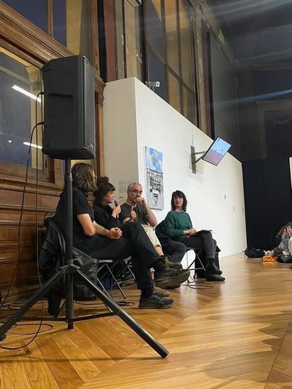 Photo en contre-plongée des quatre intervenant·es. De gauche à droite, on a Théo Pall, Margot Bernard, Vincent Romagny et Marie Preston. La rencontre se déroule dans une salle avec un beau parquet, et on voit aussi une grande cimaise blanche sur laquelle sont accrochées des oeuvres d'art.