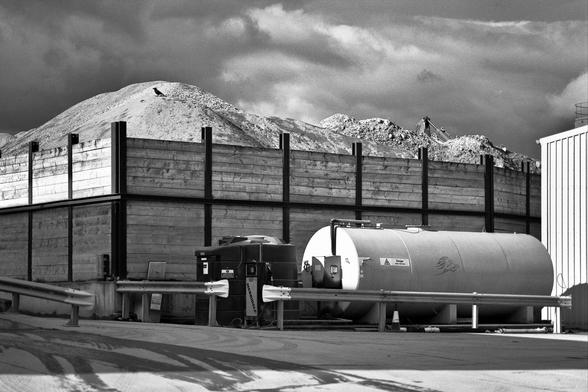 Black-and-white photograph of an aggregates works, with mounds of raw material hoppered in an enclosure, and drums in front of the enclosure.