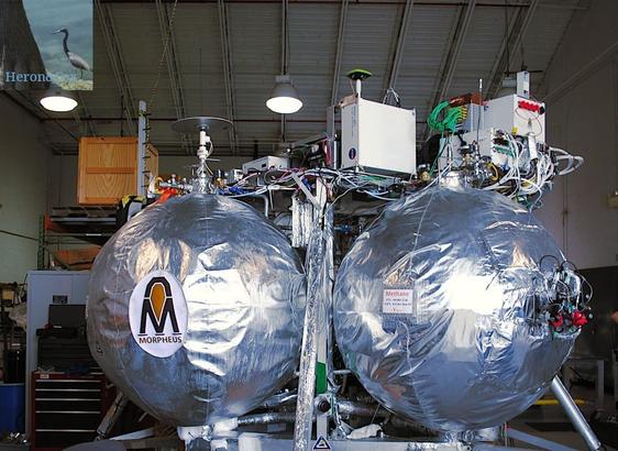 An indoor photograph of a rocket-powered lander. Two spherical tanks are in the center, covered by silvery foil. Metal struts connect the tanks and a platform on top of the tanks that is covered in electronics gear. The lander is inside a small hanger.