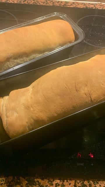 Two loafs of vegan sandwich bread still in the baking tins. The top of the loafs is golden brown. The tins are standing on a black kitchen surface to cool down.