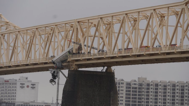Full length of a semi truck is dangling off the steel truss bridge with the cab farthest from safety and partially detached