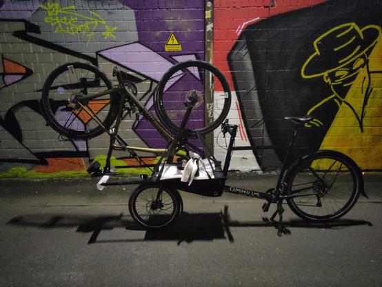Auf der Ladefläche von einem schwarzen Lastenrad steht kopfüber ein Rennrad und ist so festgezurrt.