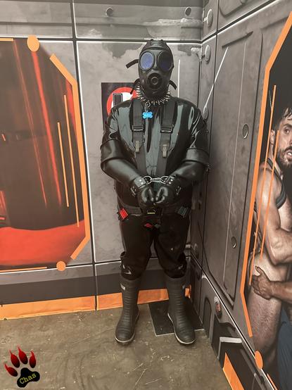 person in heavy rubber suit, covered head to toe with boots, gloves and S10 mask, hands bound, standing in a corner of a room