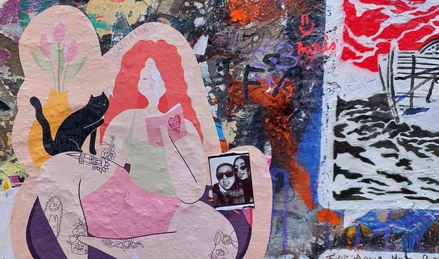 Bunt beklebte und besprühte Hauswand, auf die ein großers Papierbild geklebt ist. Eine junge Frau in Shorts und Tanktop mit wallenden roten Haaren und tätowierten Beinen liest ein Buch. Seitlich hinter ihr eine Vase mit Tulpen. Auf ihrem Bein sitzt eine schwarze Katze, die mit einer Pfote ihre Haare berührt.