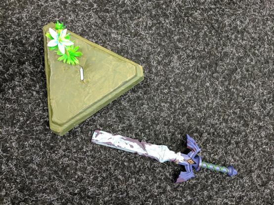 A triangular block with a flower growing from it, looking from above. In the middle is a slot where a sword would've been stuck in. The sword lays on the ground next to the block but the tip is missing, snapped off in the block