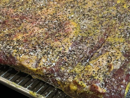 Raw brisket generously seasoned with salt, pepper, and a mustard binder on a wire rack.