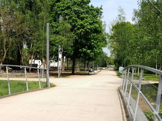 Kreuzung in einem Park, aufgenommen an einem sonnigen Sommertag, Blick von einer flachen Brücke hinein in die Kreuzungssituation. Die hinter der Brücke kreuzenden Wege sind sehr breit und aus hellem festen Sand. Am linken Weg steht an der Einbiegung zum kreuzenden Weg eine Ampel, die aus der aufgenommenen Perspektive nicht als solche gekennzeichnet ist. Am Ampelmast ist ein Anforderungskontakt. Es gibt keine weiteren Lichtzeichen, die mit der Schaltung korrespondieren.