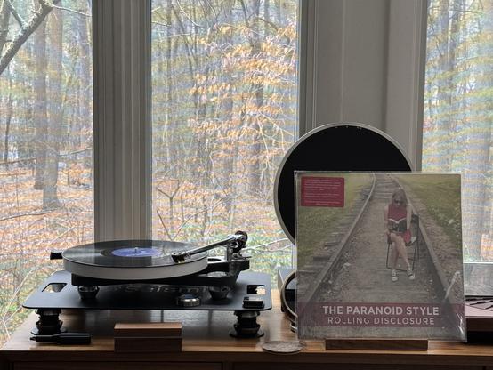 The Paranoid Style - Rolling Disclosure LP cover.

Elizabeth Nelson sitting on a folding chair in the middle of some train tracks.

The black LP plays on a Rega turntable to the left.