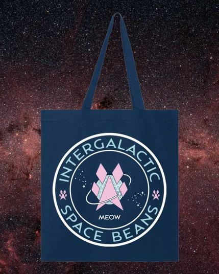 A tote bag featuring The Intergalactic Space Beans insignia - a circular logo with the words Intergalactic (top) Space Beans (bottom). A pink angular paw print in the middle against a field of white stars., encircled by a shooting star. The word MEOW where the date would be the silly human Space Force logo.