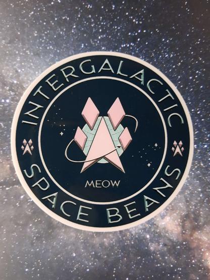 A 3" circular sticker The Intergalactic Space Beans insignia - a circular logo with the words Intergalactic (top) Space Beans (bottom). A pink angular paw print in the middle against a field of white stars., encircled by a shooting star. The word MEOW where the date would be the silly human Space Force logo.