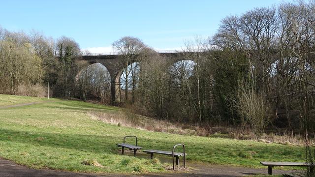 An old railway viaduct (5 of its 7 arches visible) stretching from left to right across a wooded valley with grass and benches to sit on in front. The sky behind is blue.