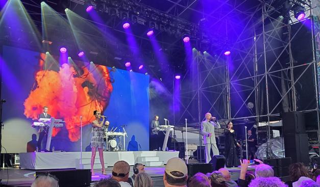 Performers on stage, lit in purple and blue, backed by a projection of a fiery coloured jellyfish. A plinth at the back has two white synths with a blonde guy on the lefthand one and a guy with dark dreadlocks on the right, with a drummer seated at an electronic drum kit between them. The three vocalists are in front: on the left, a very slim blonde woman in a sleeveless mini dress with silver fringes from  top to bottom, hair scraped back in a black scrunchie; in the centre a bald man in what looks like a light grey double breasted suit but was actually houndstooth; on the right a woman with a dark ponytail, in a long black subtly sparkling dress with a very wide deep V neckline. There are a few tops of the backs of audience members' heads in the foreground.