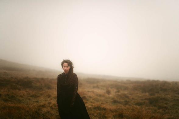 The image captures a bride in a black dress, standing in an open, foggy landscape. The dress appears long with lace sleeves, adding an element of elegance and timelessness. The bride is positioned sideways to the camera, her gaze calm and introspective, contributing to a contemplative mood. Her hair is loosely styled with strands blowing in the wind, giving the image a sense of movement and wild grace. The fog creates a mysterious ambiance and renders the landscape indistinct, drawing focus to the bride and her attire. The color palette is subdued, with earthy tones that emphasize the moody and dark atmosphere.