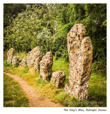 Seven stones set in grass, forming part of an ancient circle, with a worn path on the inner side, with a tall hedge behind.