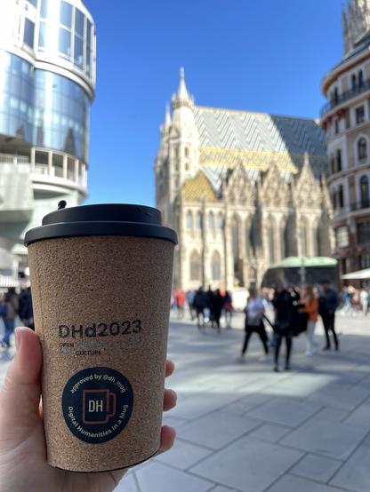 Kaffeebecher aus Kork mit DHd2023 Schriftzug und DH Mug Pickerl in einer Hand vorm Stephansdom in Wien. Im Hinterhrund sind Menschen
