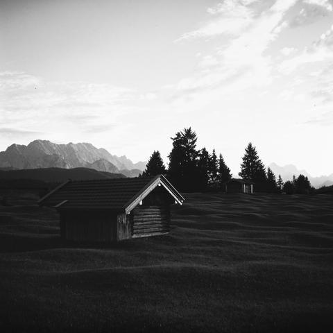 Monochrome photo of a tiny wooden hax house standing on a bumpy grassland in front of the mountains in the eavning light.