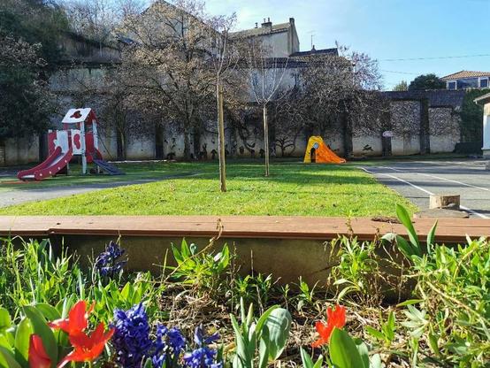 Cour de récréation végétalisée d'école maternelle, fleurs au premier plan et structures de jeu à l'arrière-plan.