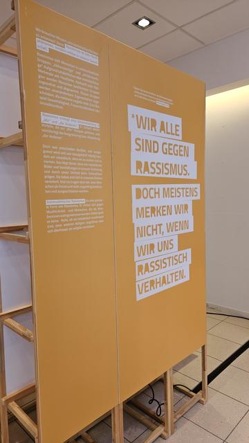 Eine Schrifttafel aus der Ausstellung. Gelbe Schrift auf weißem Grund sagt: Wir alle sind gegen Rasissmus. Doch meistens merken wir nicht, wenn wir uns rassistisch verhalten.