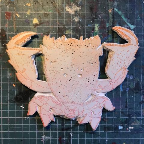 Photo of a carved lino block with orange tinge of a frog crab viewed from above. The background has been cut off the block and the crab has spots, hairs and some texture carved into it. It sits on a cutting mat with some ink stains.
