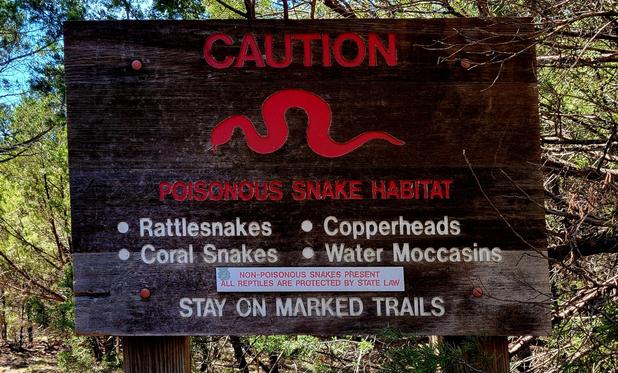 A brown wooden warning sign in a forest with red and white text, and a red silhouette of a snake:

CAUTION
[snake silhouette]
POISONOUS SNAKE HABITAT
• Rattlesnakes   • Copperheads
• Coral Snakes   • Water Moccasins

STAY ON MARKED TRAILS
